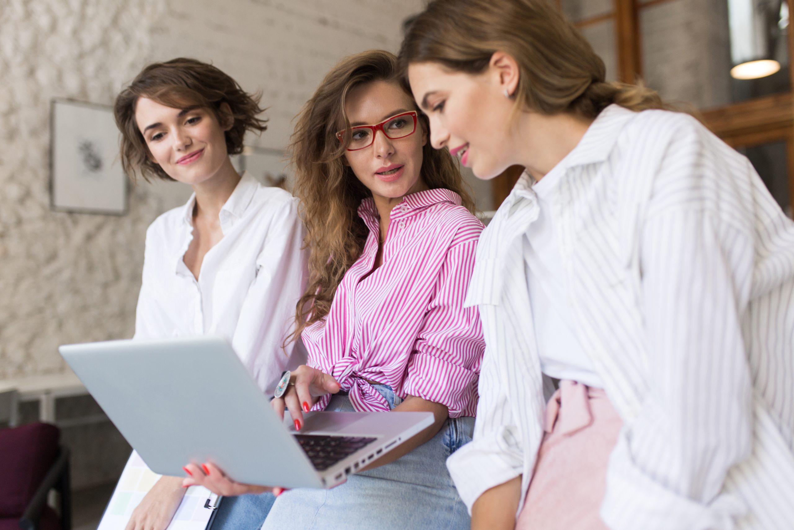 Young woman in pink shirt and eyeglasses thoughtfully discussing work with colleagues at work. Group of stylish girls spending time together in modern office